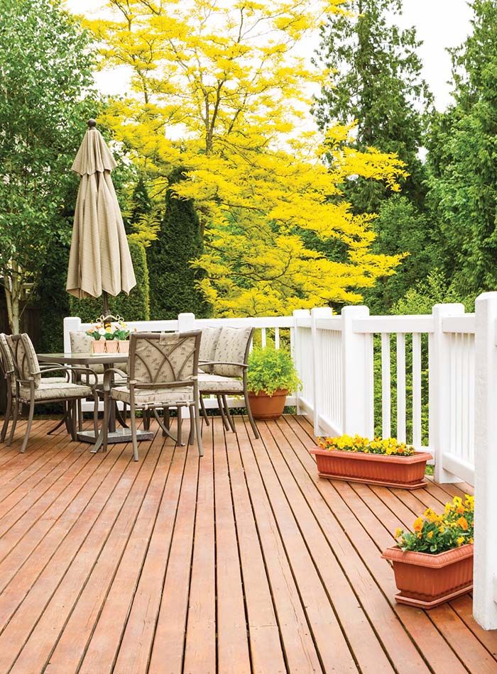 A cozy wooden deck featuring a round table and chairs, surrounded by vibrant green trees and bright yellow foliage. Flower pots line the edges.