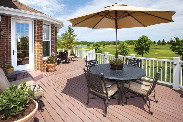 A sunny patio featuring a round table with chairs, an umbrella, and potted plants, overlooking lush green fields and trees.