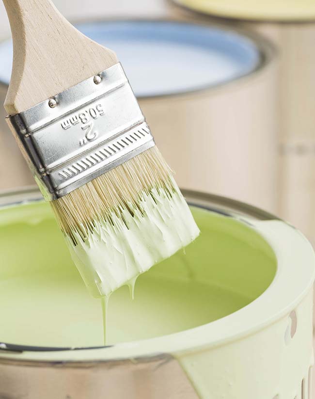 A paintbrush dipped in light green paint, dripping slightly, with paint cans of various pastel colors blurred in the background.