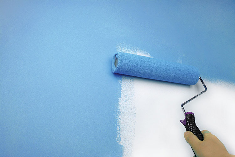 A hand using a paint roller to apply a light blue color on a wall, transitioning from a white base to a blue surface.